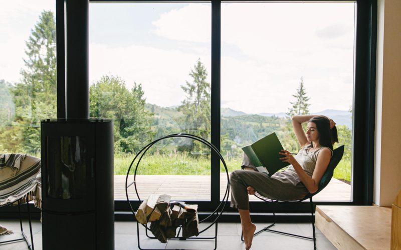 Beautiful stylish woman reading book on chair at fireplace with firewood on background of mountain hills. Young female in casual clothes relaxing in modern chalet with amazing view from window